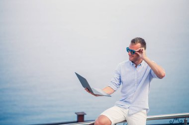 Genç başarılı erkek işadamı deniz kenarında dizüstü bilgisayarla çalışıyor. Güneş gözlüğü, gömlek ve beyaz şort giyiyor. Ofisin dışında çalış, serbest çalışan.