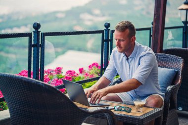 Başarılı bir erkek işadamı, dağ manzaralı bir dizüstü bilgisayarın arkasında tatilde çalışıyor. Çevrimiçi yönetici iş akışı. Balkondan güzel bir manzarayla dışarıda çalışmak.