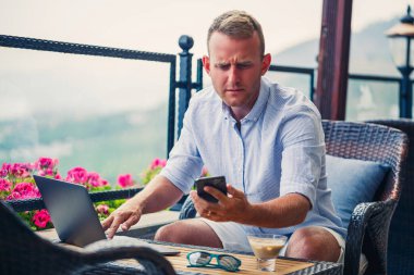Erkek iş adamı güzel panoramik manzaralı bir dizüstü bilgisayarla tatilde çalışıyor. Başarılı bir yönetici kahve içiyor ve kahve içerken telefonla konuşuyor.