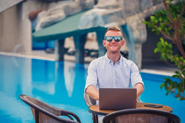 Güneş gözlüklü genç erkek işadamı havuzun yanındaki masada oturan bir dizüstü bilgisayarda çalışıyor. Uzaktan kumanda. Serbest Çalışan