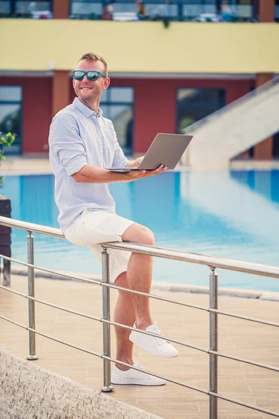 Genç erkek iş adamı, dizüstü bilgisayarı havuz kenarında oturuyor. Açık işyeri. Serbest yazar konsepti. İnternetten alışveriş. Tatil işi.