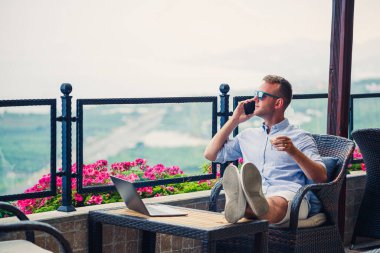 Erkek iş adamı güzel panoramik manzaralı bir dizüstü bilgisayarla tatilde çalışıyor. Başarılı bir yönetici kahve içiyor ve kahve içerken telefonla konuşuyor.