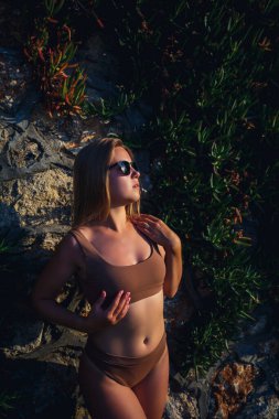 Bej mayo giymiş güneş gözlüklü güzel seksi kız. Bronzlaşmış vücutlu genç bir kadın. Taş duvarın yanında yeşil bitkilerle dikiliyor.