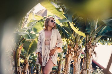 Avrupa görünümlü uzun sarı saçlı güzel genç bir kadın muz ağaçlarının yanında duruyor. Güneşli bir yaz gününde Tropikal ormanda bir kız