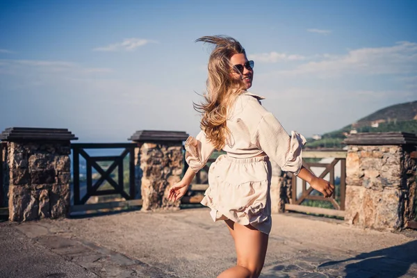 A young woman with long blond hair in a top and a skirt is walking on a summer sunny day. Happy girl with a smile on her face and in sunglasses is walking on a sunny day