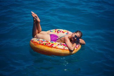 Mavi denizde gevşeyen şişme yüzüklü mayolu güzel, mutlu genç bir kadın. Güneşli bir gün, deniz kenarında tatil, turizm