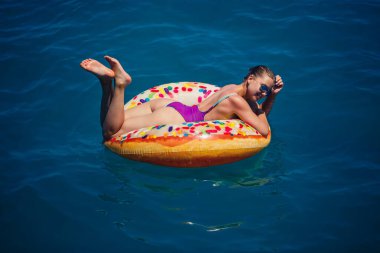 Mavi denizde gevşeyen şişme yüzüklü mayolu güzel, mutlu genç bir kadın. Güneşli bir gün, deniz kenarında tatil, turizm