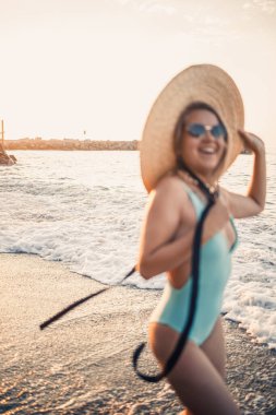 Gün batımında, mayolu ve gözlüklü şapkalı bir kadın kumsalda yürüyor. Deniz eğlencesi konsepti. Seçici odak