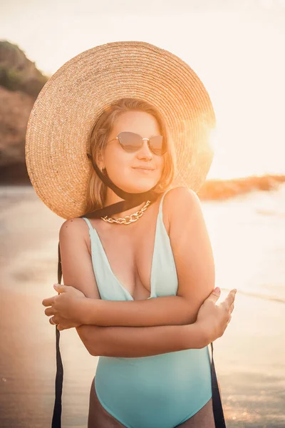 藁帽子をかぶった美しい水着の若い日焼けした女性が立っており 砂と熱帯のビーチに残り 夕日と海を見ています 選択的フォーカス 海による休暇の概念 — ストック写真