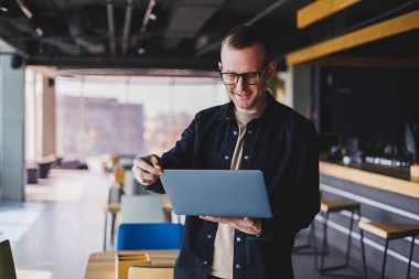 Siyah tişörtlü ve gözlüklü bir erkek girişimci elinde dizüstü bilgisayarla ofiste duruyor, başarılı şirket patronu zengin yaşam tarzından memnun.