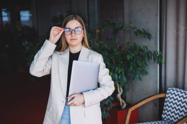 Ofiste çalışan başarılı bir yöneticinin portresi. Bilgisayarlı mutlu bir kadın modern ofiste eğleniyor. Sarı saçlı, bej ceketli ve gözlüklü bir kadın.