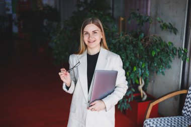 Ofiste çalışan başarılı bir yöneticinin portresi. Bilgisayarlı mutlu bir kadın modern ofiste eğleniyor. Sarı saçlı, bej ceketli ve gözlüklü bir kadın.