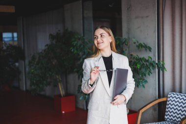 Ofiste çalışan başarılı bir yöneticinin portresi. Bilgisayarlı mutlu bir kadın modern ofiste eğleniyor. Sarı saçlı, bej ceketli ve gözlüklü bir kadın.