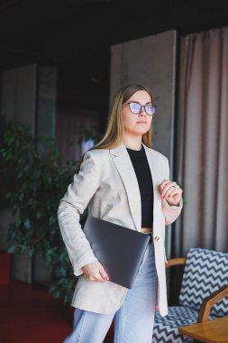 Ofiste çalışan başarılı bir yöneticinin portresi. Bilgisayarlı mutlu bir kadın modern ofiste eğleniyor. Sarı saçlı, bej ceketli ve gözlüklü bir kadın.