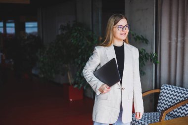 Ofiste çalışan başarılı bir yöneticinin portresi. Bilgisayarlı mutlu bir kadın modern ofiste eğleniyor. Sarı saçlı, bej ceketli ve gözlüklü bir kadın.