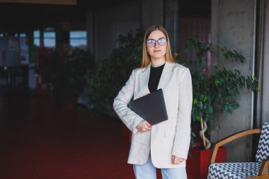 Ofiste çalışan başarılı bir yöneticinin portresi. Bilgisayarlı mutlu bir kadın modern ofiste eğleniyor. Sarı saçlı, bej ceketli ve gözlüklü bir kadın.