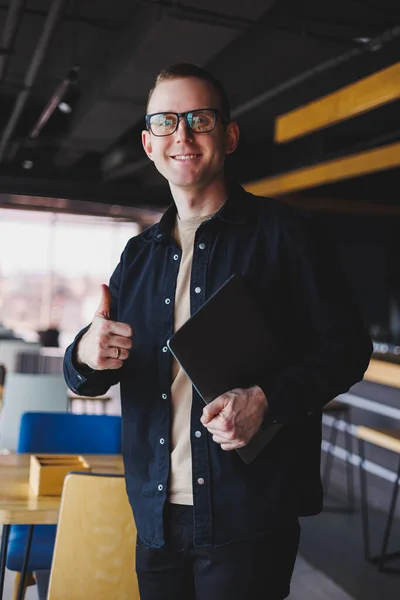 Male entrepreneur in black shirt and glasses standing in office with laptop in hand, successful corporate boss feeling good from rich lifestyle