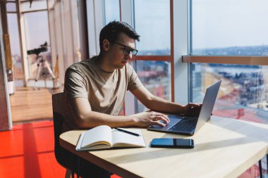 Genç milenyum erkeği çevrimiçi çalışırken dizüstü bilgisayarın önünde oturuyor. Modern dijital göçebe çalışmaları, kafenin iç kısımlarında netbook 'ta serbest çalışma üzerine