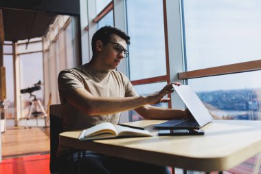 Genç milenyum erkeği çevrimiçi çalışırken dizüstü bilgisayarın önünde oturuyor. Modern dijital göçebe çalışmaları, kafenin iç kısımlarında netbook 'ta serbest çalışma üzerine