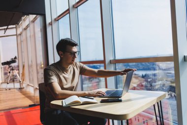 Genç milenyum erkeği çevrimiçi çalışırken dizüstü bilgisayarın önünde oturuyor. Modern dijital göçebe çalışmaları, kafenin iç kısımlarında netbook 'ta serbest çalışma üzerine