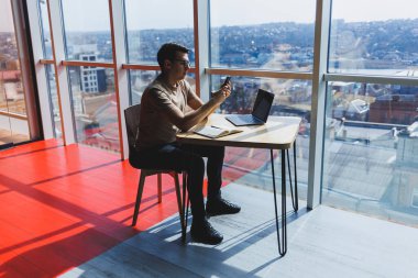 Kırmızı tişörtlü yakışıklı adam, kafeteryada uzaktan çalışan bir dizüstü bilgisayarla cam pencerenin yanında otururken cep telefonuna mesaj yazıyor.