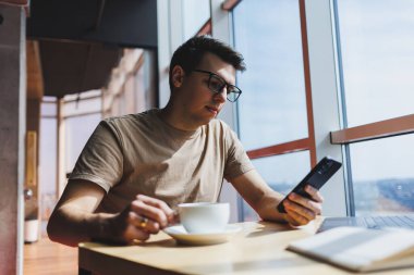 Kırmızı tişörtlü yakışıklı adam, kafeteryada uzaktan çalışan bir dizüstü bilgisayarla cam pencerenin yanında otururken cep telefonuna mesaj yazıyor.