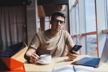 Kırmızı tişörtlü yakışıklı adam, kafeteryada uzaktan çalışan bir dizüstü bilgisayarla cam pencerenin yanında otururken cep telefonuna mesaj yazıyor.