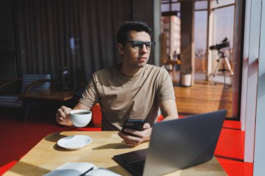 Kırmızı tişörtlü yakışıklı adam, kafeteryada uzaktan çalışan bir dizüstü bilgisayarla cam pencerenin yanında otururken cep telefonuna mesaj yazıyor.