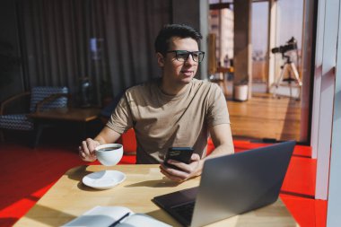 Kırmızı tişörtlü yakışıklı adam, kafeteryada uzaktan çalışan bir dizüstü bilgisayarla cam pencerenin yanında otururken cep telefonuna mesaj yazıyor.