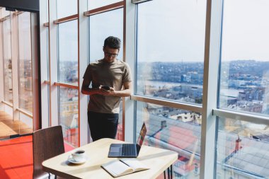Neşeli Scofe erkek girişimci ofis penceresinin yanında duruyor ve şirkette çalışırken gülümsüyor başarılı şirket patronu zengin yaşam tarzından memnun.