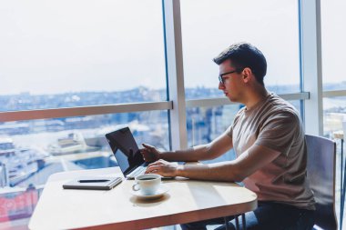 Genç milenyum erkeği çevrimiçi çalışırken dizüstü bilgisayarın önünde oturuyor. Modern dijital göçebe çalışmaları, kafenin iç kısımlarında netbook 'ta serbest çalışma üzerine