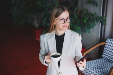 Akıllı telefondan konuşan ve kafede dikilirken kahve içen mutlu bir kadın. Bir kafede duran gülümseyen sarışın kadın. Dinlenmek ve boş zaman kavramı. Modern kadın yaşam tarzı