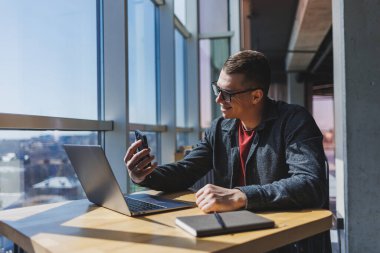 Gözlüklü neşeli serbest çalışan akıllı telefonlu ve gülümseyen gün boyunca kafede dizüstü bilgisayarlı ve not defterli bir masada oturuyor. Ofisten uzakta çalışıyor..