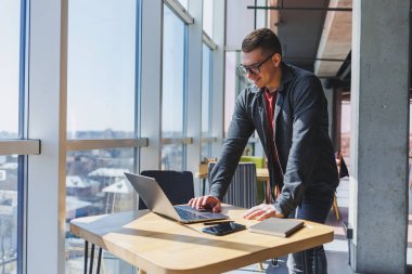 Modern bir dizüstü bilgisayarla uzaktan çalışan, mola sırasında masaya oturup kameraya gülümseyen, vizyon düzeltme gözlüklü mutlu bir insan programcı portresi.