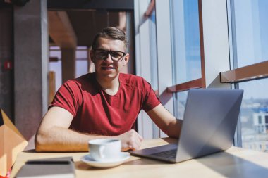 Modern bir dizüstü bilgisayarla uzaktan çalışan, mola sırasında masaya oturup kameraya gülümseyen, vizyon düzeltme gözlüklü mutlu bir insan programcı portresi.
