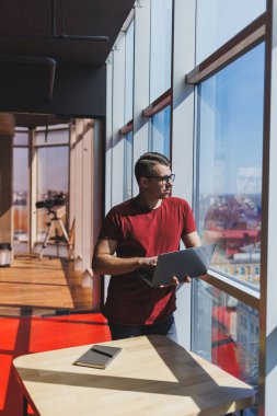 Gülümseyen işadamı bir dizüstü bilgisayar üzerinde çalışırken pencerenin yanında duruyor. Çoklu görev konsepti. Gözlüklü meşgul bir serbest yazar pencerenin yanında dururken elinde bir dizüstü bilgisayar tutuyor. Akıllı gündelik.