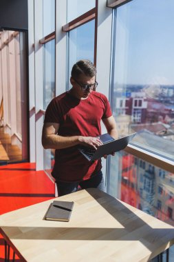 Gülümseyen işadamı bir dizüstü bilgisayar üzerinde çalışırken pencerenin yanında duruyor. Çoklu görev konsepti. Gözlüklü meşgul bir serbest yazar pencerenin yanında dururken elinde bir dizüstü bilgisayar tutuyor. Akıllı gündelik.