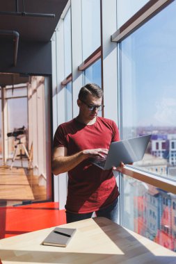 Gülümseyen işadamı bir dizüstü bilgisayar üzerinde çalışırken pencerenin yanında duruyor. Çoklu görev konsepti. Gözlüklü meşgul bir serbest yazar pencerenin yanında dururken elinde bir dizüstü bilgisayar tutuyor. Akıllı gündelik.