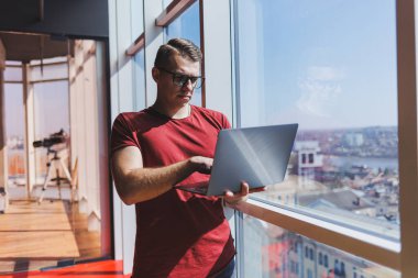 Gülümseyen işadamı bir dizüstü bilgisayar üzerinde çalışırken pencerenin yanında duruyor. Çoklu görev konsepti. Gözlüklü meşgul bir serbest yazar pencerenin yanında dururken elinde bir dizüstü bilgisayar tutuyor. Akıllı gündelik.