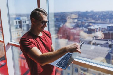 Gülümseyen işadamı bir dizüstü bilgisayar üzerinde çalışırken pencerenin yanında duruyor. Çoklu görev konsepti. Gözlüklü meşgul bir serbest yazar pencerenin yanında dururken elinde bir dizüstü bilgisayar tutuyor. Akıllı gündelik.