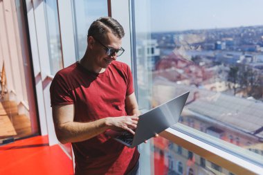 Gülümseyen işadamı bir dizüstü bilgisayar üzerinde çalışırken pencerenin yanında duruyor. Çoklu görev konsepti. Gözlüklü meşgul bir serbest yazar pencerenin yanında dururken elinde bir dizüstü bilgisayar tutuyor. Akıllı gündelik.