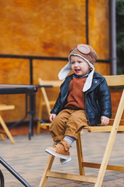 Üç yaşında, deri ceketli, kahverengi pantolonlu şık bir çocuk kafenin yanındaki terasta oturuyor. Modern çocuk. Çocuk modası. Mutlu çocuk