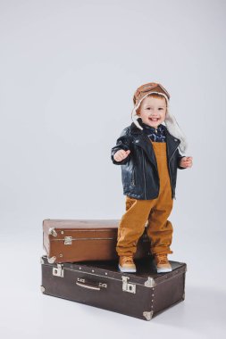 Deri ceketli ve kahverengi tulumlu küçük bir çocuk büyük bir bavulun üzerinde duruyor. Bavullu bir çocuk. Küçük gezgin..