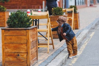 Üç yaşında, deri ceketli, kahverengi pantolonlu şık bir çocuk kafenin yanındaki terasta oturuyor. Modern çocuk. Çocuk modası. Mutlu çocuk