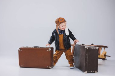 Kasklı ve pilot ceketli mutlu bir çocuk kahverengi bir bavul taşıyor. Bir çocuk pilotun portresi, deri ceketli bir çocuk. Bavullu küçük gezgin.
