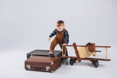 Kahverengi tulumlu neşeli bir çocuk bavulun üzerinde duruyor. Bavullu küçük bir gezgin. Tahta uçak, çocuk oyuncakları.