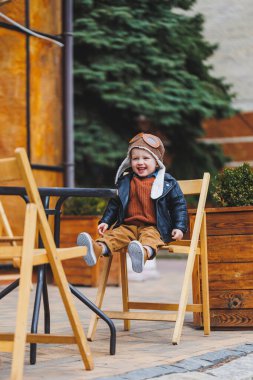 Üç yaşında, deri ceketli, kahverengi pantolonlu şık bir çocuk kafenin yanındaki terasta oturuyor. Modern çocuk. Çocuk modası. Mutlu çocuk