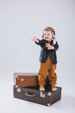 Deri ceketli ve kahverengi tulumlu küçük bir çocuk büyük bir bavulun üzerinde duruyor. Bavullu bir çocuk. Küçük gezgin..