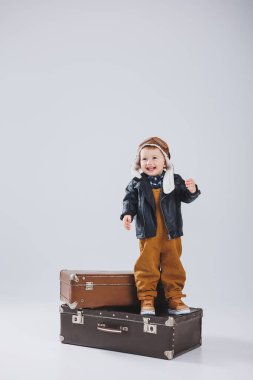 Deri ceketli ve kahverengi tulumlu küçük bir çocuk büyük bir bavulun üzerinde duruyor. Bavullu bir çocuk. Küçük gezgin..
