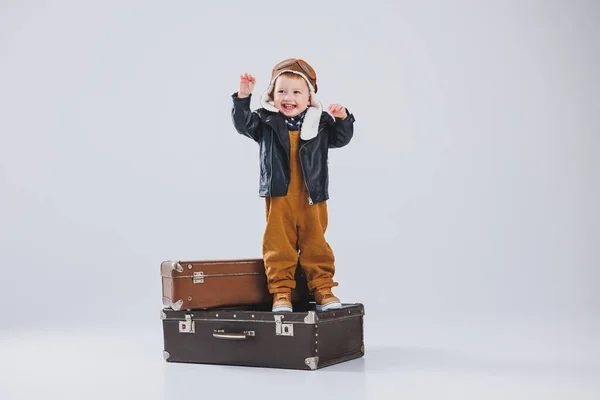 A small boy in a leather jacket and brown overalls stands on a large suitcase. Child with a suitcase. Little traveller.